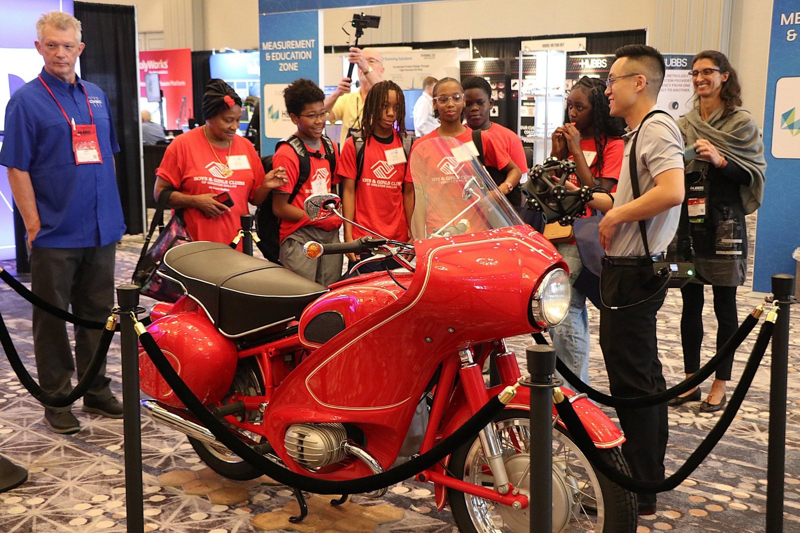Students from the Boys & Girls Clubs of Greater Dallas watching a handheld 3D scanner measure a red vintage motorcycle at the CMSC Education Zone Students from the Boys & Girls Clubs of Greater Dallas watching a handheld 3D scanner measure a red vintage motorcycle at the CMSC Education Zone