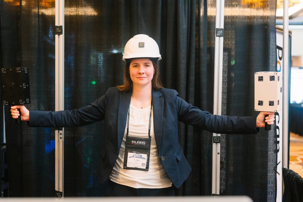 Conference attendee in a hard hat and CMSC badge standing between two measurement stands with arms outstretched