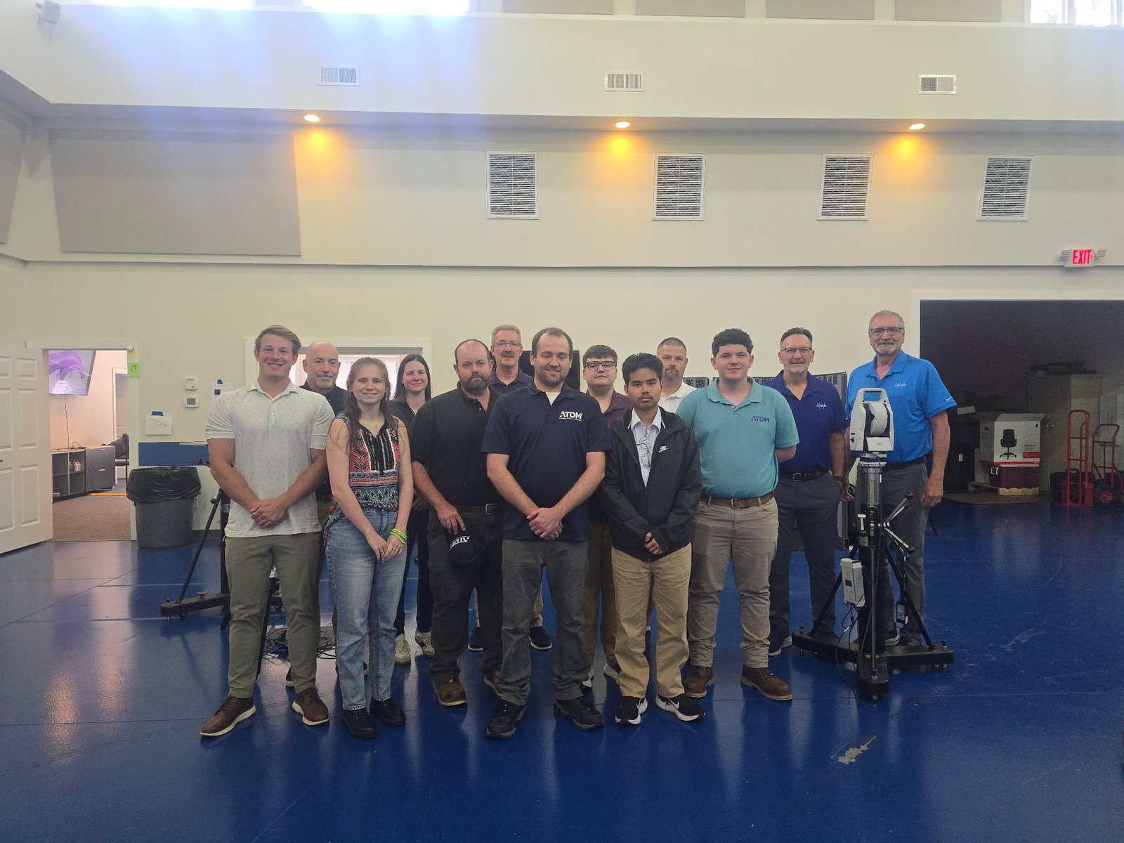 Group of metrology training class participants standing together beside a laser tracker on a tripod in a training room